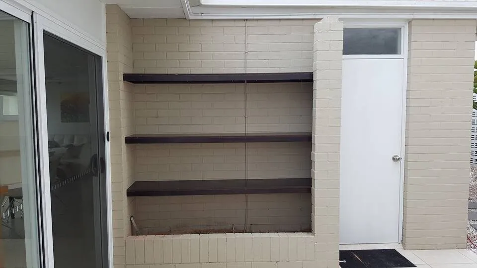 Three dark timber shelves fixed across a painted brick alcove beside the back door.