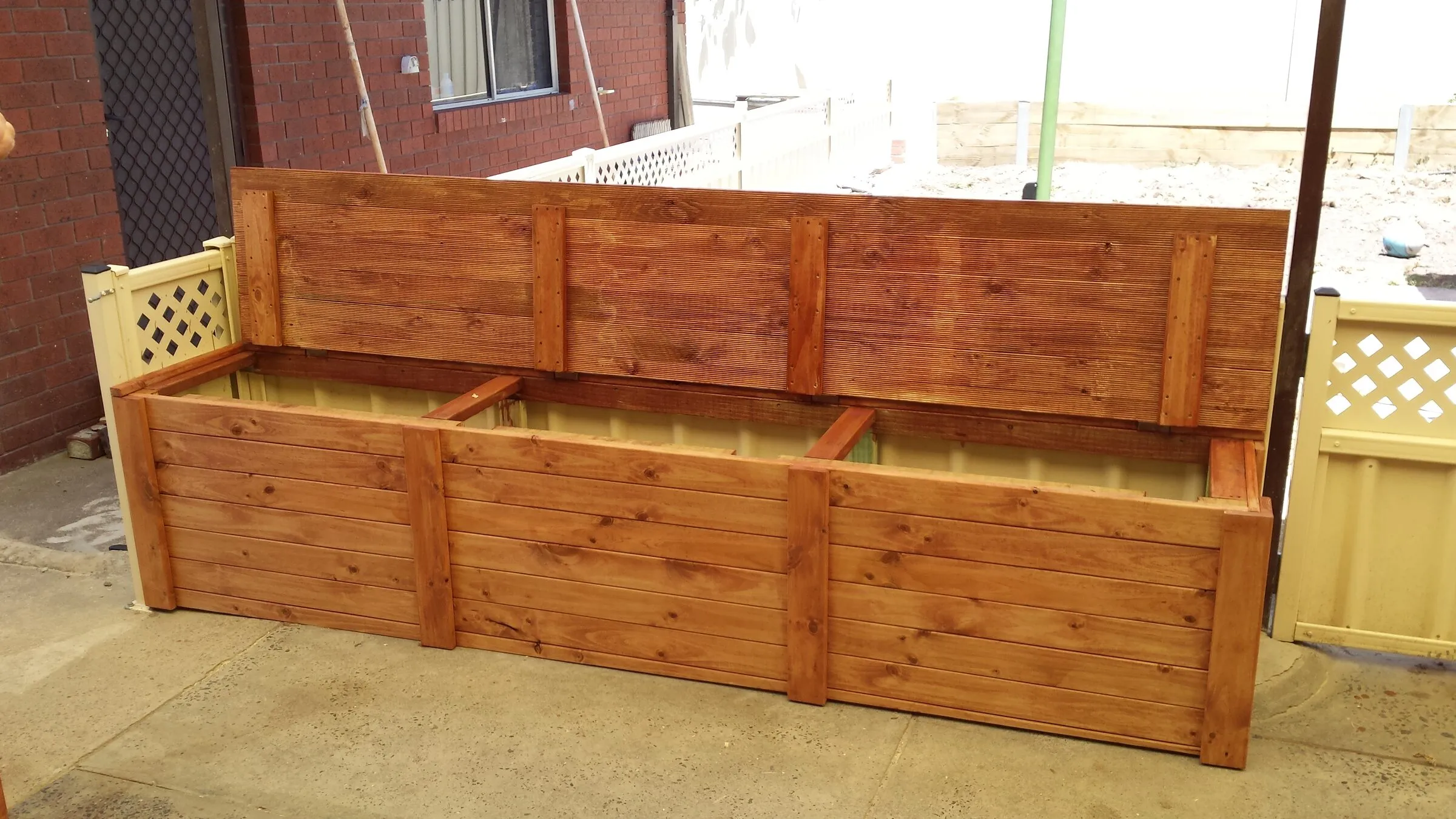 A long timber storage bench with lift-up lids, built and stained against a brick wall.