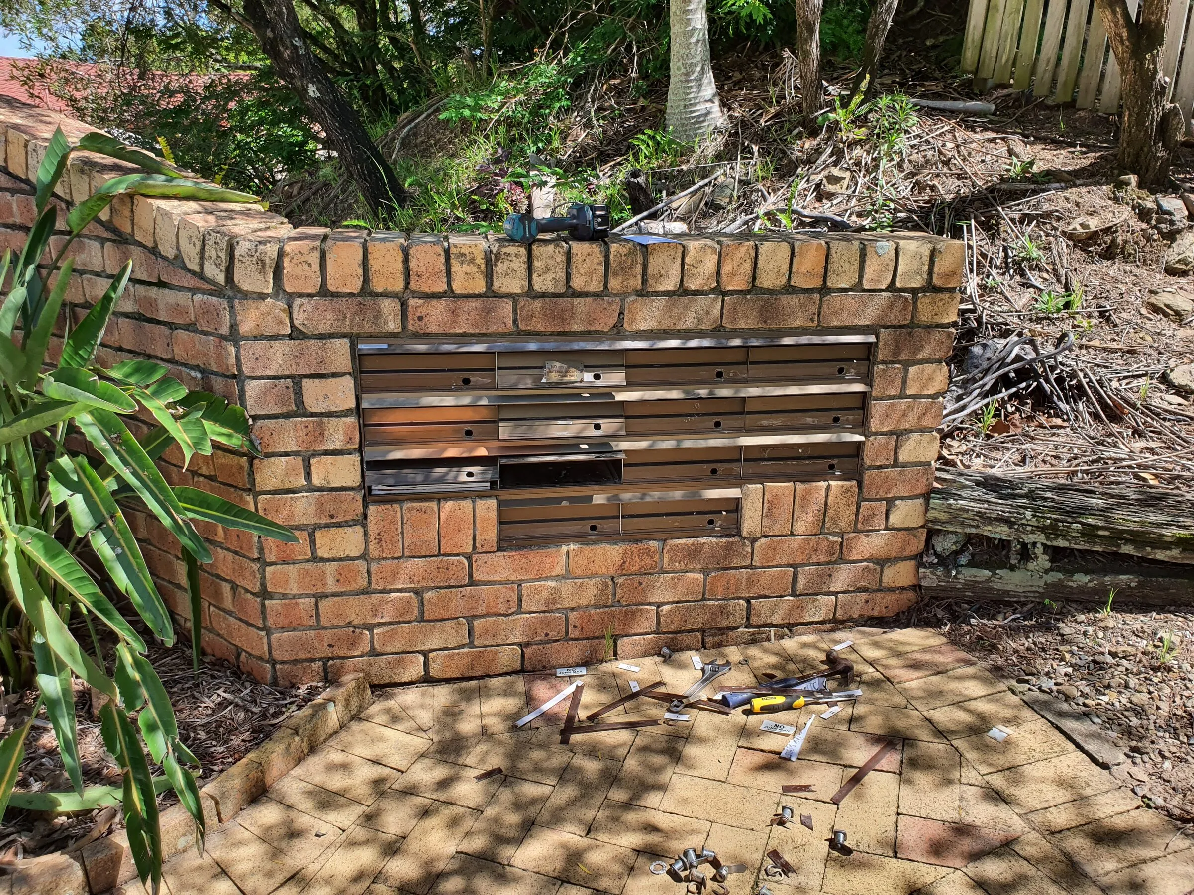 The old stainless letterbox bank coming out of the brick wall, fixings and tools laid out on the pavers.