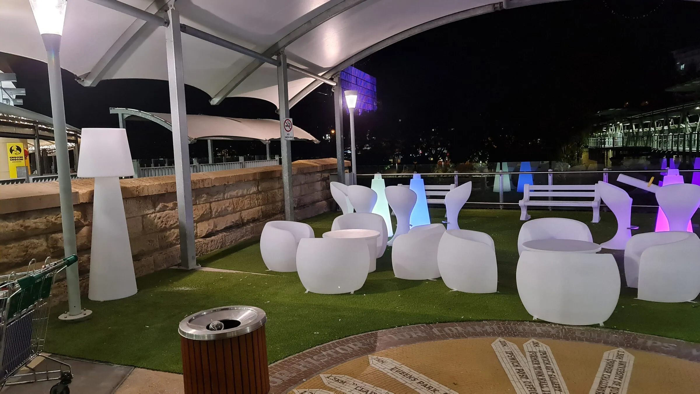 Illuminated outdoor lounge chairs and tables set out on the turf under a shade canopy.